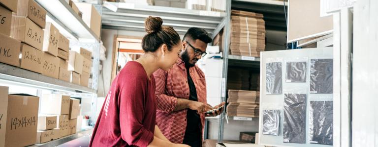 A man and a woman look at a phone together in a warehouse surrounded by cardboard boxes