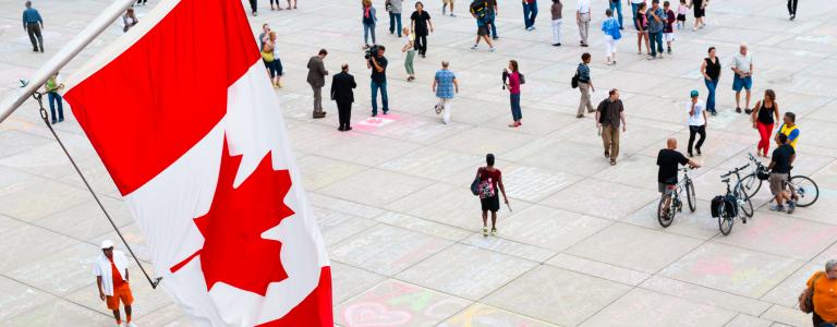 A crowd seen from above mills about a town square with the Canadian flag hanging on the left