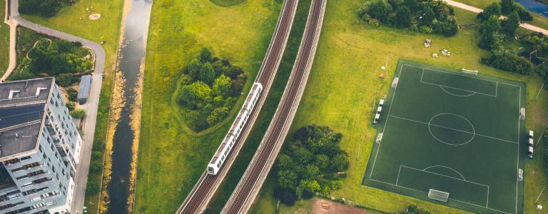 Aerial view of a train going down a track, surrounded by green fields and buildings