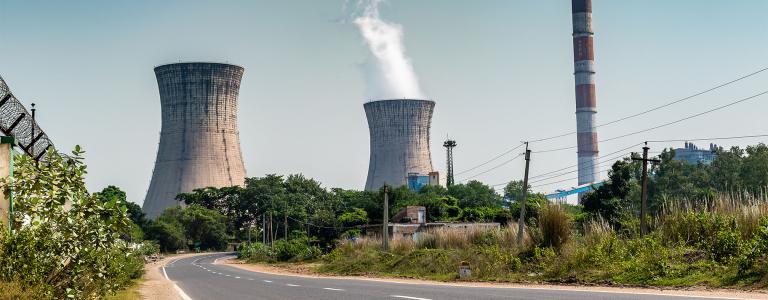 A road leads towards the towers of a coal power station. Smoke emerges from one of the towers set against a blue sky.