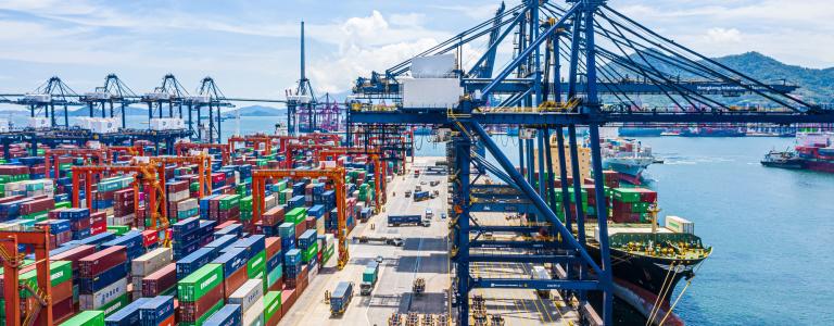 Aerial view of a shipyard with many stacked cargo containers on the left and ships in the harbour on the right.