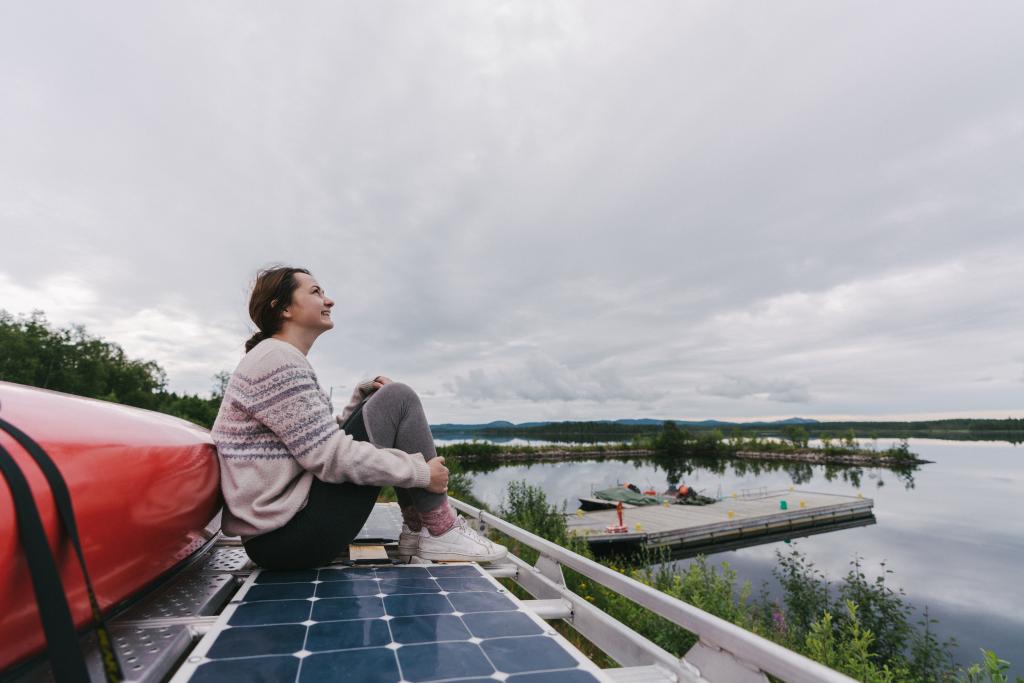 A young woman wearing a sweater sits on top of a camper van next to solar panels looking into an overcast sky and smiling